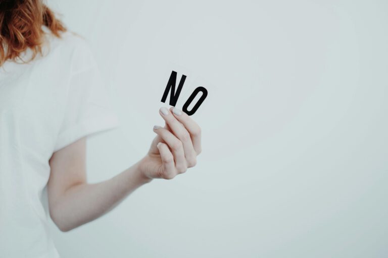 Frau in weißem T-Shirt steht vor einer weißen Wand und hält das Wort NO in ihren Händen. Dieses Foto symbolisiert das Setzen von eigenen Grenzen.
