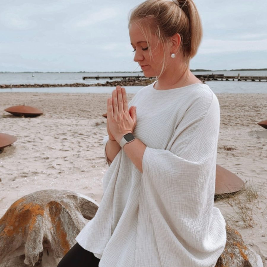 Sophia meditiert am Strand und hält ihre Hände in Gebetshaltung vor ihrem Herzen zusammen als Zeichen der Ruhe, inneren Mitte und Entspannung