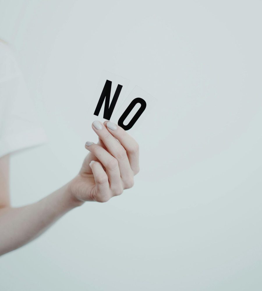 Frau in weißem T-Shirt steht vor einer weißen Wand und hält das Wort NO in ihren Händen. Dieses Foto symbolisiert das Setzen von eigenen Grenzen.